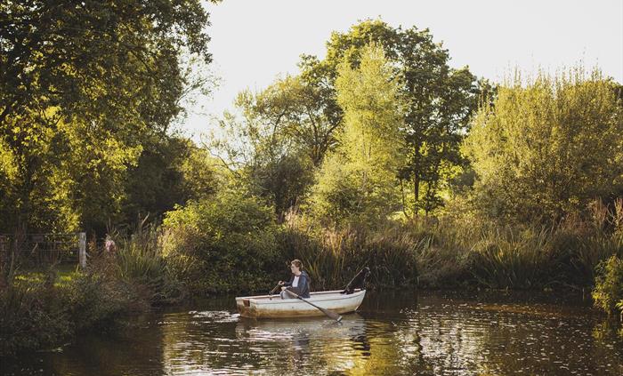 A girl in a rowing boat on a pond with a dog looking out.